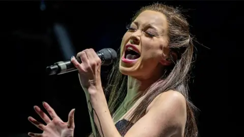 Elliott Davis A woman with dark hair on a stage with her mouth open as she belts a song. She holds a microphone close to her face.