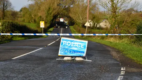 A two lane country road with hedgerows and green fields either side. A road closed police sign is on the road and a barrier is across the road. A back van can be seen at the bottom of the road.