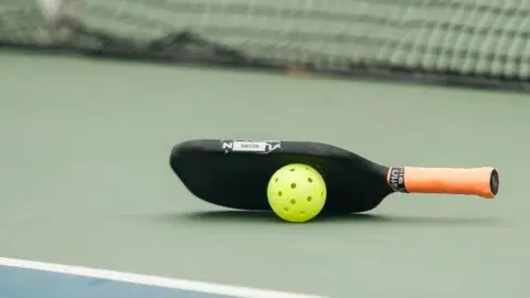 Getty Images A racquet and ball used in pickleball placed on the surface of a court. There is a net in the background.