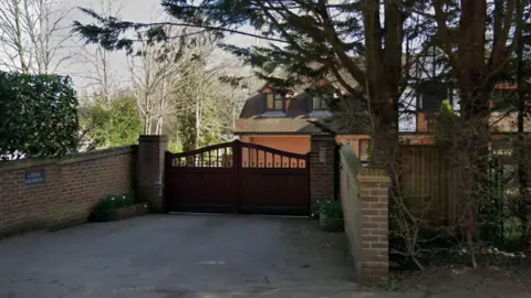 The image shows the outside of a house, where there is a large gate in-between brick walls and shrubbery. In the background another house can be seen.