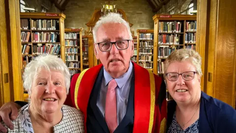 Noel standing in the Bangor library with his university robes, flanked by his daughter and wife