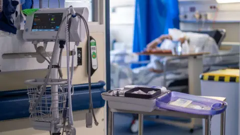 Hospital equipment in a hallway near the entrance of a ward. A patient laying down on a bed with their arm going towards their head can be seen in the blurred background.