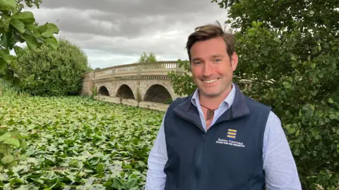 A man with short brown hair in a side parting, who is wearing a blue gilet over a checked white and blue shirt, stands in front of a lake covered with water lilies. A stone bridge stands in the background. Three arches are visible along with a decorative balustrade.