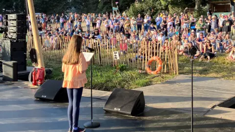 Jon Wright/BBC A girl stands on stage with a crowd in front watching her speak into a microphone.