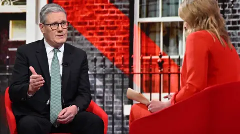 Prime Minister Sir Keir Starmer in the Sunday with Laura Kuenssberg studio. He wears a black suit, white shirt and mint green tie and sits opposite Kuenssberg, who is wearing a red trouser suit.