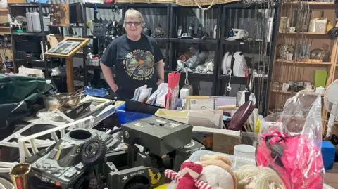 A middle-aged woman with short cropped hair and glasses stands with her hands in her pockets, she is smiling as she is surrounded by bric-a-brac.