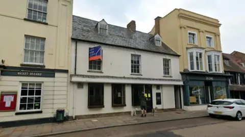 Google The former Lloyds Bank in Towcester. It is a two-storey white building with a slanted slate roof. A man walking two dogs is looking into the window.