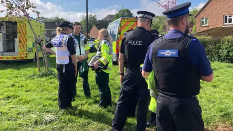 A number of police and PCSOs are standing on a patch of grass in the middle of houses, where a police van and ambulance are parked.
