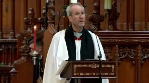 Rev Canon Alan Bashforth is standing at a lectern in a church setting. He is wearing vestments. 