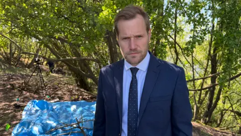 BBC/Jim Scott Det Supt Matt Steel is standing in front of a tree unsmiling. He has blond hair and a slight beard. He is wearing a blue shirt and a navy tie and suit jacket. There is a large blue plastic sheet on the ground behind him. 