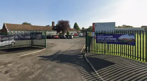 A single-storey school building seen from a green entrance gate into the car park with a sign saying Higham lane School and a fabric banner advertising a sixth form open evening. Cars are parked in front of the building and a grassy area is seen to the right of the gate. It is a sunny day and the sky is blue with a few clouds.