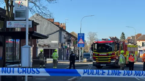 Police cordon in place with a 'Police Line Do Not Cross' tape is pictured. In the background, a Leicestershire Fire and Rescue Service crew is working on Saffron Lane in Leicester, dealing with debris from a commercial building