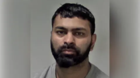 Custody image of Aamar Mohammed, a man with black hair and beard in a grey jumper is looking at the camera and against a grey background