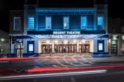 The outside of a red-brick theatre with the words Regent Theatre written on the side of a canopy above the main entrance. The photo is taken in darkness with the lights shining 