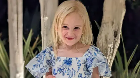 Sofia Hill. She is a young child with long blonde hair. She is wearing a blue and white floral dress and is looking at the camera and smiling.