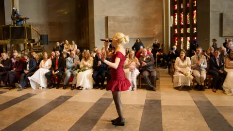 Singer in a red dress performs in the central aisle, watched by guests at Coventry Cathedral