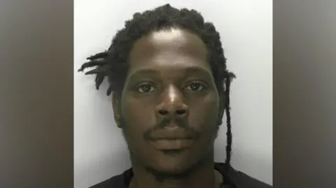 A mugshot of a man who has black dreadlocks and is wearing a black T-shirt. He is stood against a white wall and has a blank expression.
