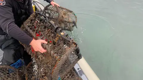 Clean Planet UK Two hands holding a lobster pot over the edge of the sea on a boat. The pot is large and brown. 