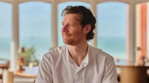 John Hersey Jamie Park is tuned away from the camera, smiling. He is sat in a restaurant with a sea view and he is wearing his chef whites. He has a ginger beard and blue eyes, and his hair is brown and curly.