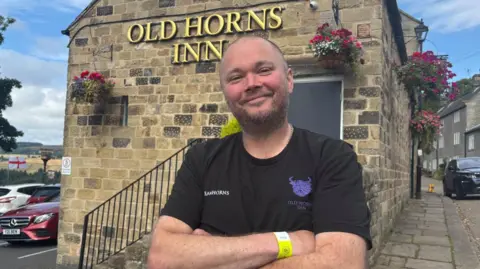 Simon Thake A man with shaved hair and a black t-shirt stands smiling outside a pub