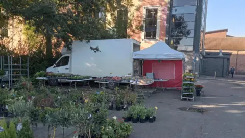 Chris Ford A white van with an awning at the back in a car park next to trees and a building. Rows of plants are dotted in front of the van