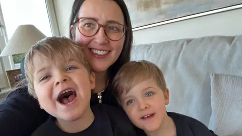 Kim Borthwick with her four-year-old twin sons Max and Freddie. She has long, dark hair and glasses and the boys have blond hair and blue eyes. They are sitting on a grey sofa in a living room and are all smiling at the camera 
