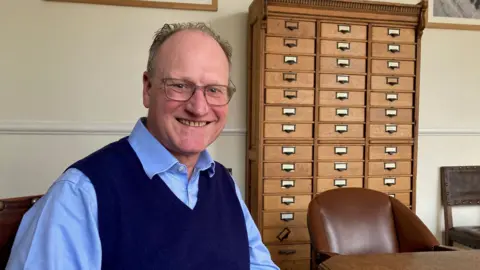 A balding man with glasses wearing a dark blue V-neck pullover and light blue shirt.