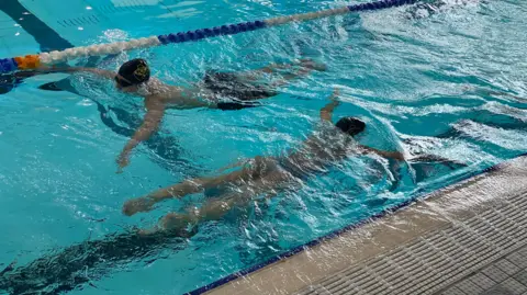 Both Gemma and Luca are in the water in the pool and the camera is over the top of them looking down. They're both doing breast stroke. Luca is wearing black swimming trunks and Gemma is in a light blue and patterned swim costume.