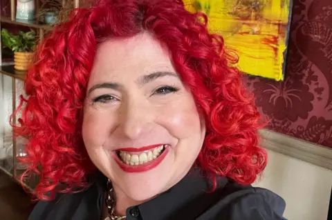 Dr Lori Beth Bisbey Dr Lori Beth grins at the camera. She has bright red curly hair and is wearing matching red lipstick. Behind her is a room with patterned wallpaper, shelving, plants, and a large rectangular artwork featuring yellow and red colours.