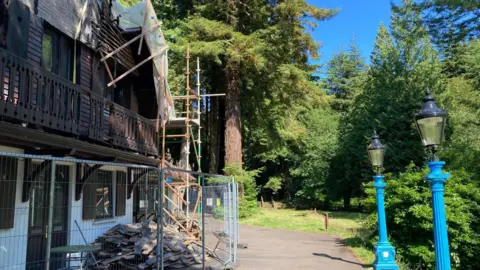 A panelled building on the left has scaffolding and fencing around it and piles of building materials. To the right are two blue traditional lamp posts with the green glen behind, with grass and tall trees on a sunny day.