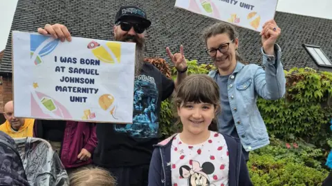Helen Campbell A family posing for a photo in the rain, a mum, dad and child smiling at the camera. The parents are holding signs in front of the child that say "I was born at Samuel Johnson maternity unit"