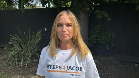 A woman with blonde hair looks at the camera, wearing a white t-shirt which says '#Steps4Jacob' and '#10forken' on it.