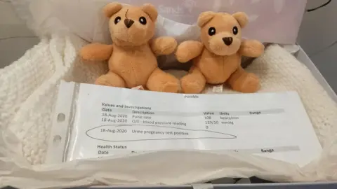 Two teddy bears and a white baby blanket are displayed in a memory box with a print out showing a positive pregnancy test result