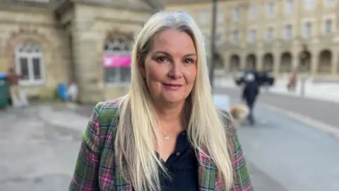 BBC Lindsay Rae who has long blonde hair and is wearing a silver necklace, black top and pink and green checked jacket 