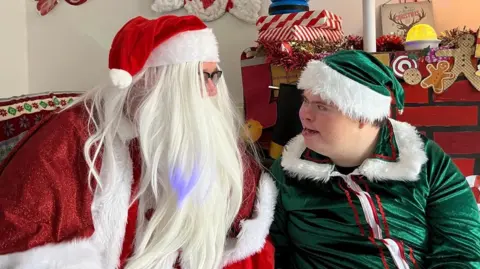 Alice Cullinane/BBC Santa Claus is wearing a red hat and a cape. He has long white hair and is sitting next to a red brick fireplace. A man with a green hat and a green suit is sitting next to Santa and smiling.