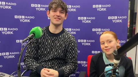 A 20-year-old man and 13-year-old girl sit in front of microphones in a radio studio, smiling at the camera.