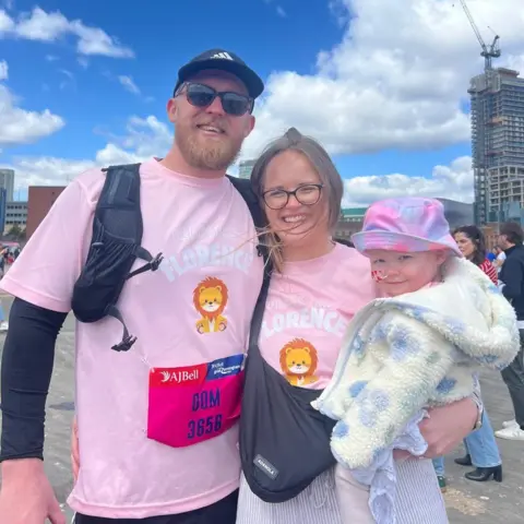 Family A man and woman smile at the camera, with the woman holding her toddler in her arms. The parents wear light pink t-shirts which say 'Fearless Florence' on them. The little girl has a nasal tube and wears a fluffy white coat with blue flowers, and a tie-dye hat.