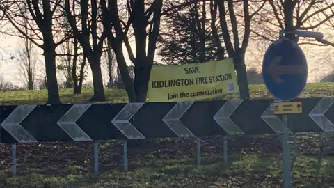 A yellow banner reading "Save Kidlington Fire Station. Join the conversation" put between the trees at the mount at Kidlington roundabout. A QR code can be seen as part of it as well.