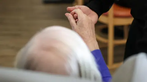 Reuters A carer holding the hand of a person in a care home seen from behind with grey hair