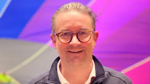 Hardy wearing round tortoiseshell glasses with his dark blond hair slicked back and smiling at the camera. He is wearing an off-white shirt with a grey jacket over the top. A rainbow hot air balloon is behind him. 