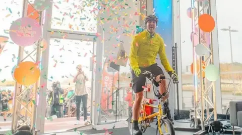 Paddy McGuiness in fluorescent cycling gear surrounded by confetti as he crosses the finish line on a kids' bike