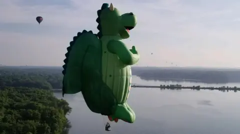International Bristol Balloon Fiesta A huge balloon in the shape of a green dragon flies over a river in a hazy day, with smaller balloons flying in the background