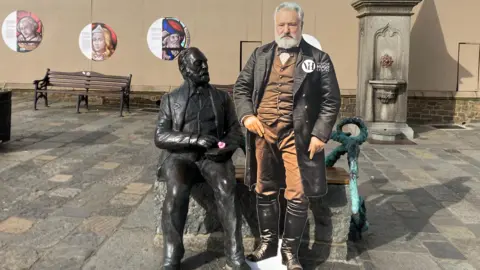 A cardboard cut out of author Victor Hugo standing next to a statue of Victor Hugo. There are benches in the background 