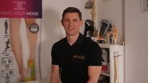 Jamie Niblock/ BBC Jimmy Reynolds, wearing a black polo top, is sitting on a treatment bed. In the background, there are models of joints and a spinal column. There are posters about the body on the wall.