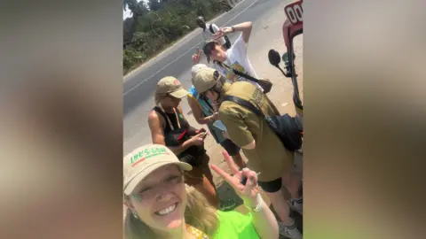 Amanda Hitchin A group of women. One is holding the camera and taking a selfie while holding up the peace sign.
She is wearing a green top and a sun hat and smiling at the camera.
There are other women in the photo some of them are talking to one another and another woman is also looking at the camera and holding up two peace signs.
