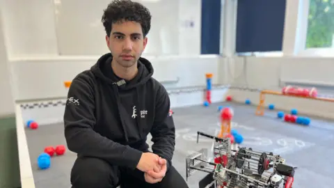 Adil is kneeling on the floor with his robot next to him. He has black curly hair and is wearing a black hoodie and black tracksuit bottoms. 