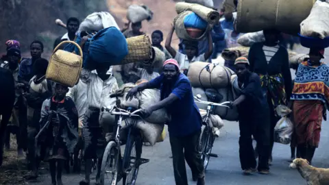 Getty Images About a dozen Rwandan Tutsi people are pushing bikes loaded with belongings or balancing loads on their heads and carrying bundles down a road. A dog is in the right hand corner of the photograph
