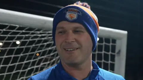 Jamie Niblock/BBC Chris Jackson is wearing a woollen blue and yellow hat with the Sands United crest. He is smiling and standing in front of the goal.