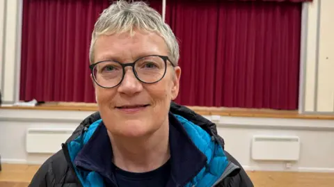 Ollie Rawlinson/BBC Linda Scott smiling into the camera. She has short, blonde hair and is wearing blue round glasses. She is standing in a village hall with a stage behind her, covered by a red curtain. She is wearing a dark padded coat with a blue hood. There is a man behind her slightly out of focus and leaning on the stage.