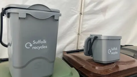 Vikki Irwin/BBC A large grey food caddy and smaller grey food bin sit on top of the largest waste bins. The words "Suffolk Recycles" are printed on their fronts. They each have a black handle. 